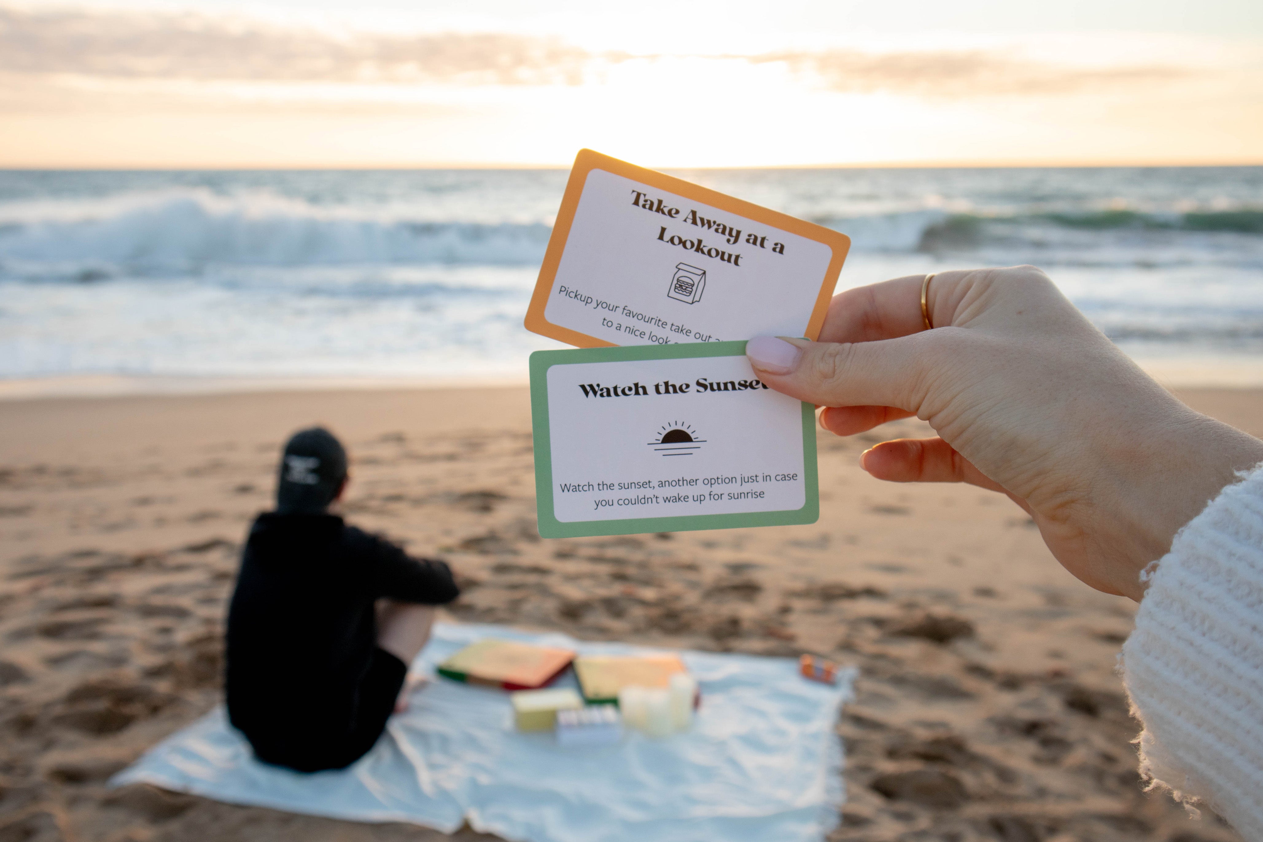 Girl showing date idea cards, date night idea cards, things to do when you're in a relationship, fun couple activities, sunset date, couple watching the sunset at the beach, couple eating pizza at the beach, pizza date, outdoor date, romantic sunset date for couples, takeaway at a lookout, affordable date night, date date ideas, set of date idea cards